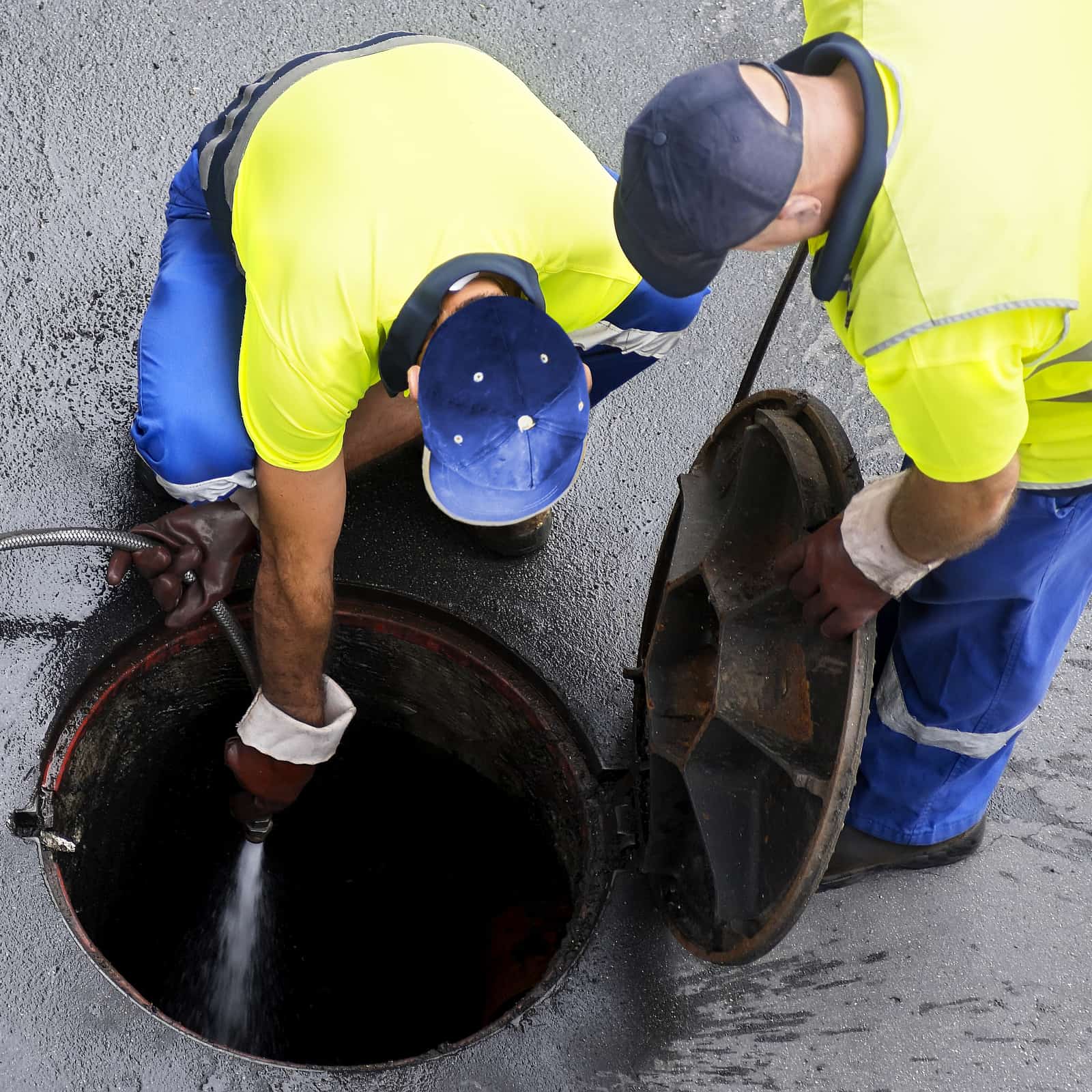 Two workers in bright yellow shirts and blue uniforms are cleaning a manhole, showcasing their expertise in cesspool services. One sprays water inside while the other holds the lid open. They remain focused on the task at hand, ensuring efficiency on the paved surface.