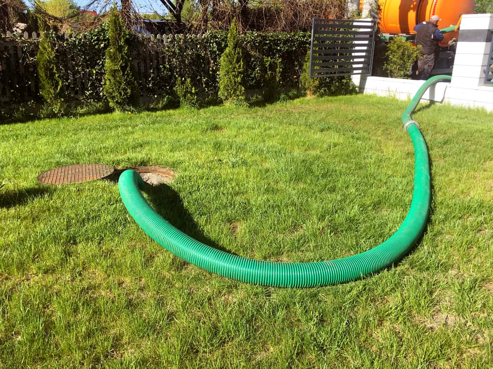 A large green hose is stretched across a grassy lawn, connected to a sewer manhole, hinting at ongoing septic tank services. The hose leads to an unseen truck off-image, with a person in the background near a white wall with a black gate.