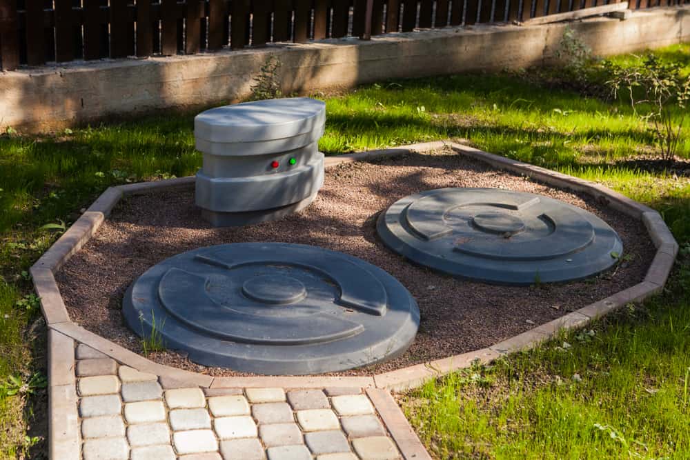 A grassy yard features two large, round gray septic tank covers, perfectly maintained by expert Septic Tank Services. Behind them sits a cylindrical piece with two buttons. The area is elegantly bordered by stones and paving stones, creating a neat stone-covered landscape.