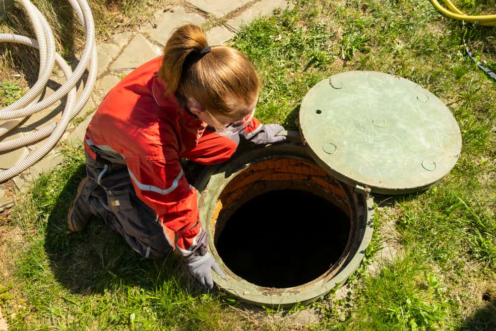 A person in safety gear inspects an open septic tank in a grassy area. They are kneeling beside the circular opening, surrounded by hoses, while performing essential septic tank services. The tank lid is partially open, indicating thorough maintenance underway.