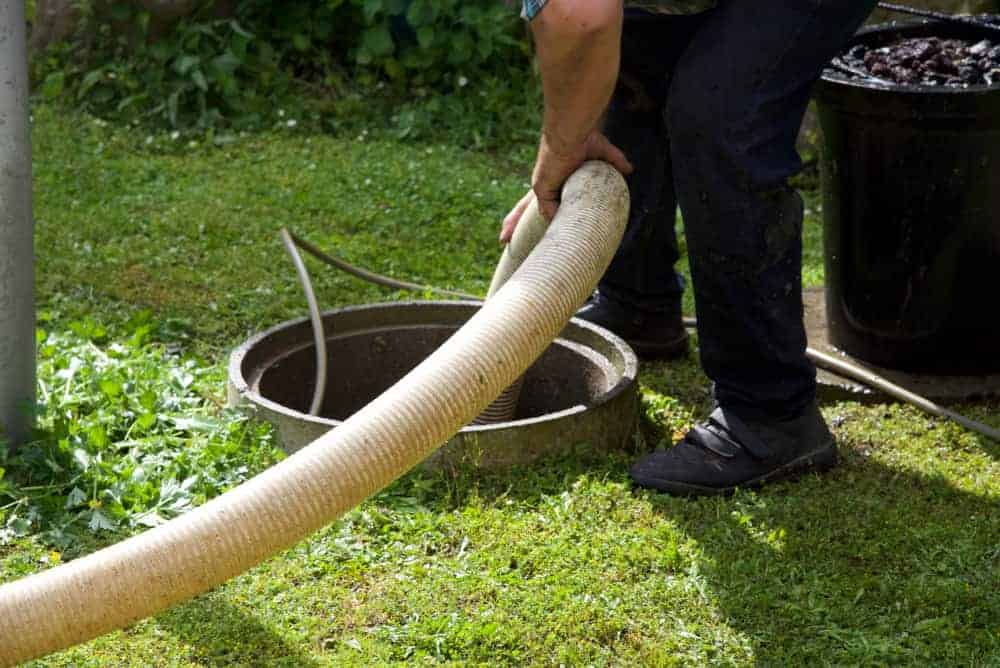 A person in dark pants and shoes is holding a large hose and inserting it into an outdoor septic tank opening surrounded by grass—showcasing professional septic tank services Long Island, NY. Another hose and a black container are nearby.