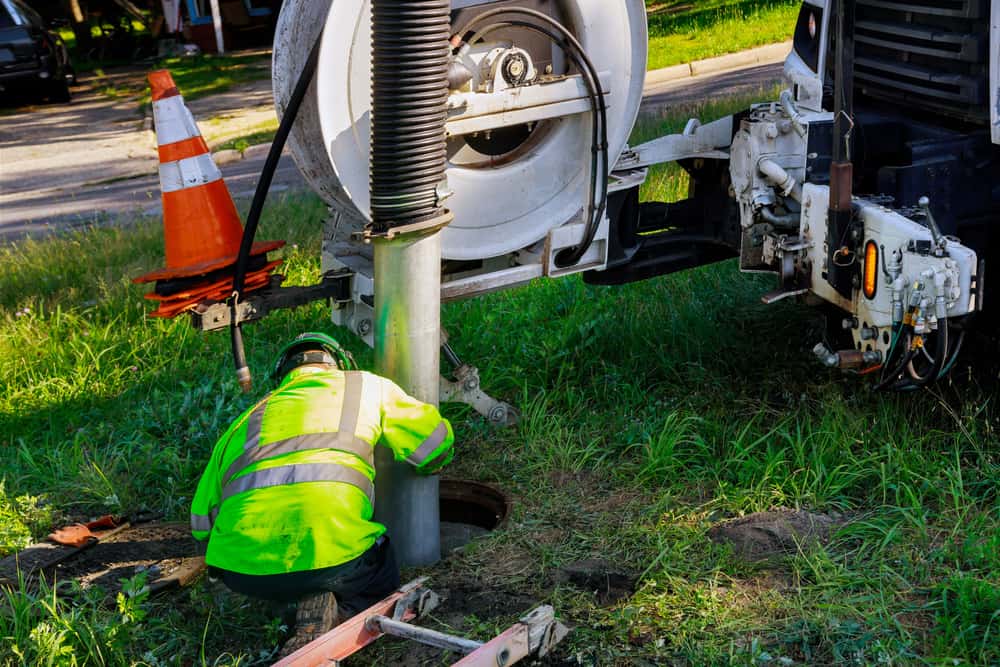 A worker in a fluorescent yellow safety jacket and helmet is operating a large vacuum truck near a manhole on grassy ground, providing cesspool services. An orange traffic cone is placed nearby as part of the safety setup.