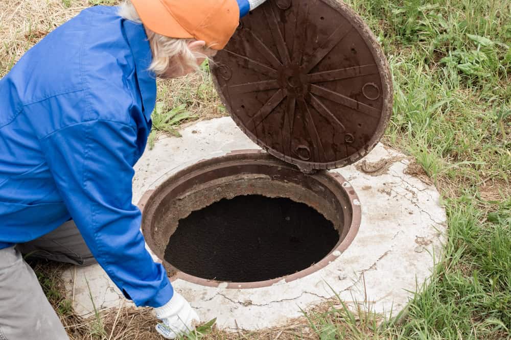 A person in a blue jacket and orange cap lifts the cover of a round septic tank on a grassy area. The open tank, maintained through professional Septic Tank Services, is visible, surrounded by concrete.