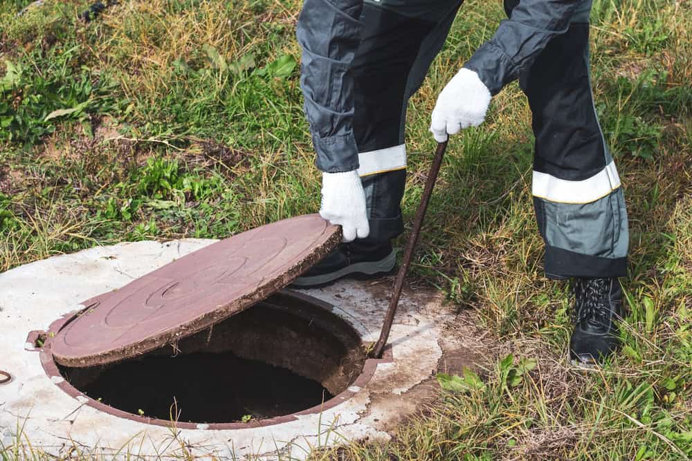 A person wearing gloves, black pants, and black boots is using a metal tool to access a circular manhole cover on a grassy area. This could be part of septic tank services as grass and weeds surround the manhole.