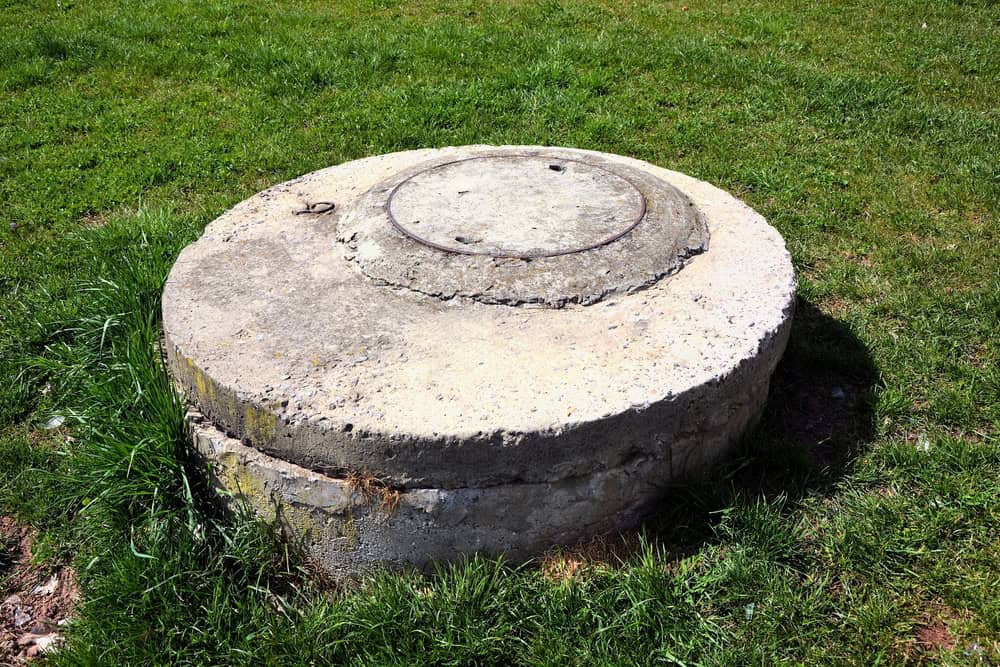 A weathered concrete manhole cover with a circular metal lid sits in the middle of a grassy area, frequently inspected by Septic Tank Services. The lush green grass surrounds the structure under a clear sky.