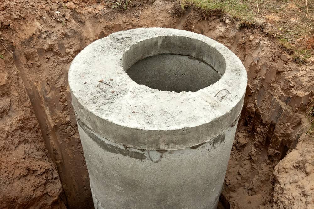 Concrete cylindrical well casing partially buried in the ground, surrounded by soil, with visible rebar loops on top. Ideal for sites requiring septic tank services due to its robust construction and easy maintenance.
