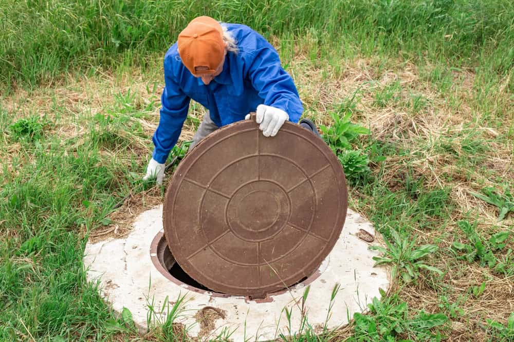 A person wearing a blue jacket, orange cap, and white gloves is carefully lifting a circular manhole cover in a grassy area, likely for septic tank services.