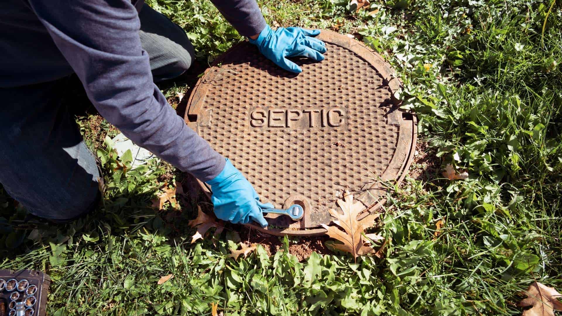 A person wearing blue gloves is kneeling on grass, using a wrench to work on a metal cover marked "SEPTIC," while leaves scatter the ground. This scene unfolds as part of comprehensive septic tank services.