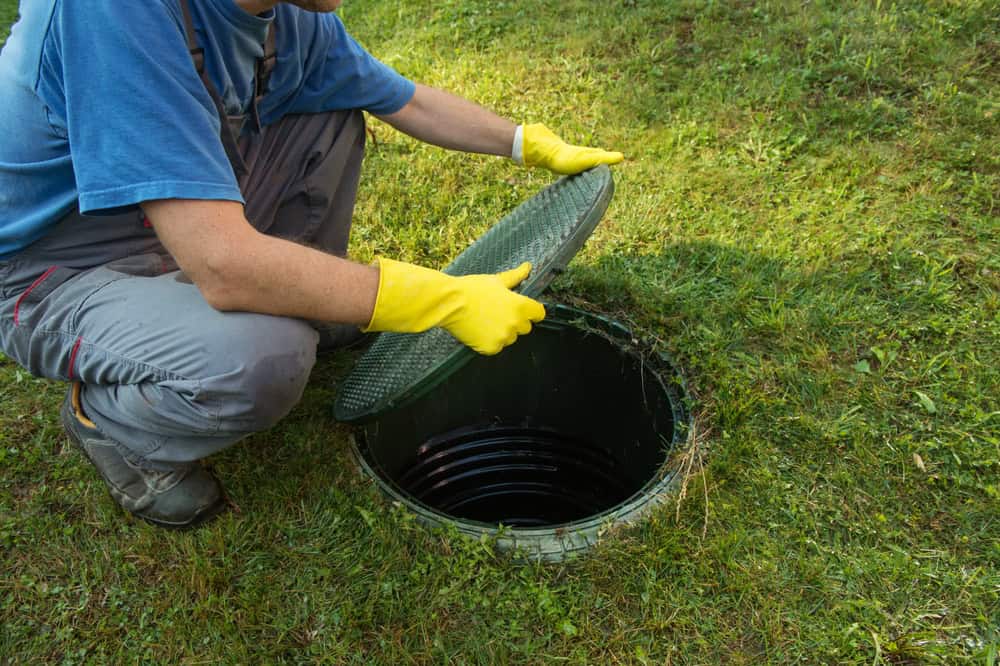 A person in a blue shirt, gray pants, and yellow gloves kneels on grass, lifting the lid of a round septic tank. This hands-on approach to septic tank services reveals the opening partially, surrounded by lush greenery.