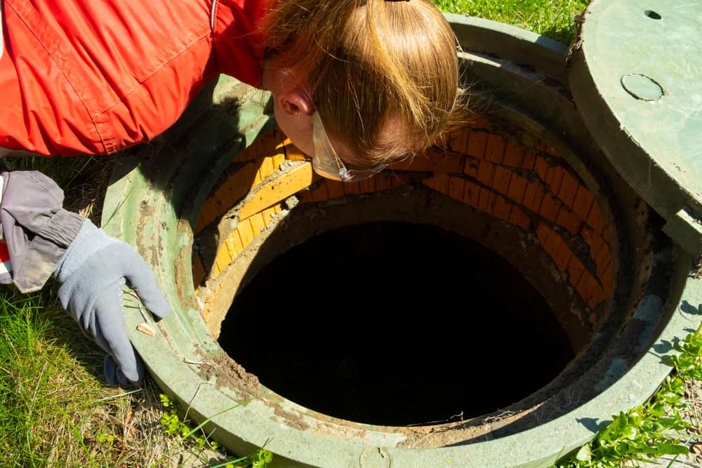 A person wearing a red jacket, gloves, and safety goggles is inspecting an open manhole, potentially part of local septic tank services. The brick-lined interior catches their focus as they lean over, peering into the dark opening amidst grass and sunlight.