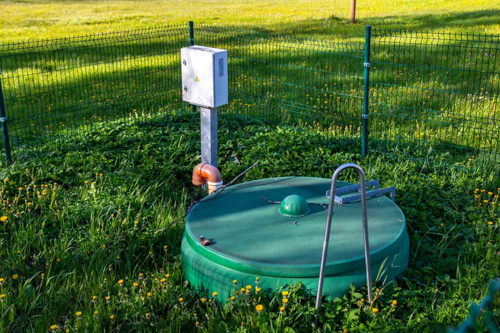 A green septic tank cover with a handle rests in a grassy field surrounded by short fencing. Nearby, a control box and a small pipe with an orange cap stand ready for septic tank services, while yellow flowers are scattered across the grass.