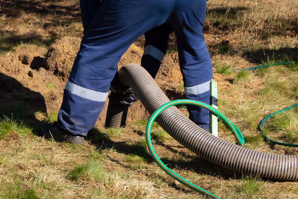 A person in dark blue work pants with reflective stripes is operating a large hose, inserting it into the ground for septic tank services in an outdoor grassy area.