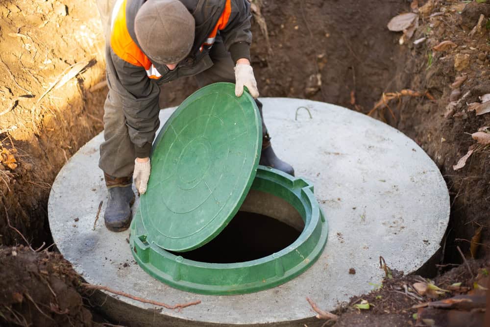 A worker wearing a cap and gloves carefully lifts a green circular lid on a concrete septic tank outdoors, demonstrating the precision of Septic Tank Services. The surrounding earthy terrain is partially adorned with fallen leaves.