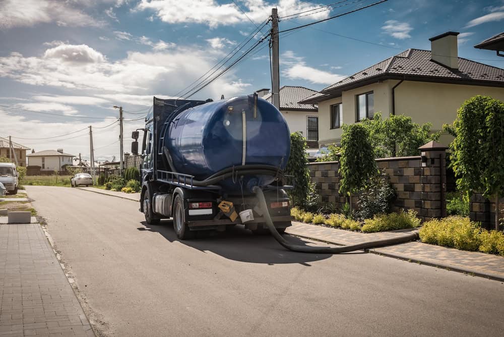 A blue sewage truck, providing septic tank services, is parked on a residential street lined with houses and gardens. A hose extends from the truck to the curb. The partly cloudy sky casts gentle shadows on the scene below.