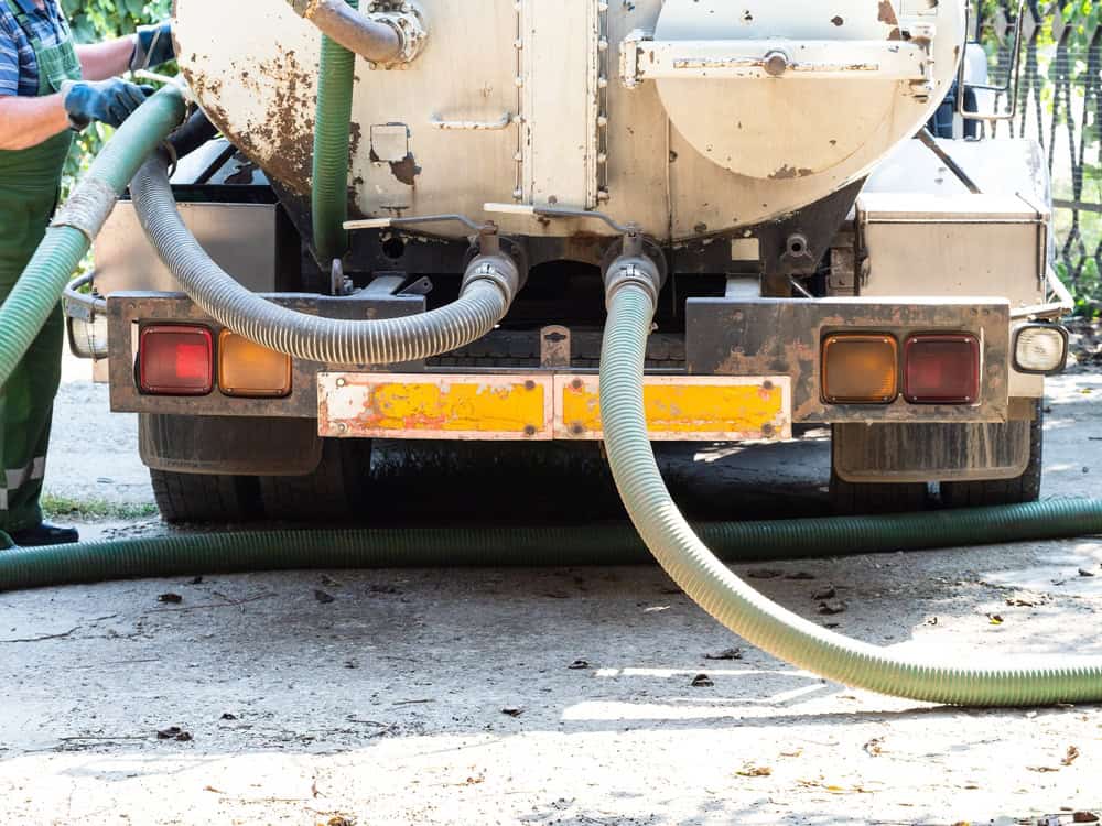A sanitation truck offering septic tank services is parked on a concrete surface, with hoses connected to its rear. A person in green pants is partially visible on the left, adjusting one of the hoses.