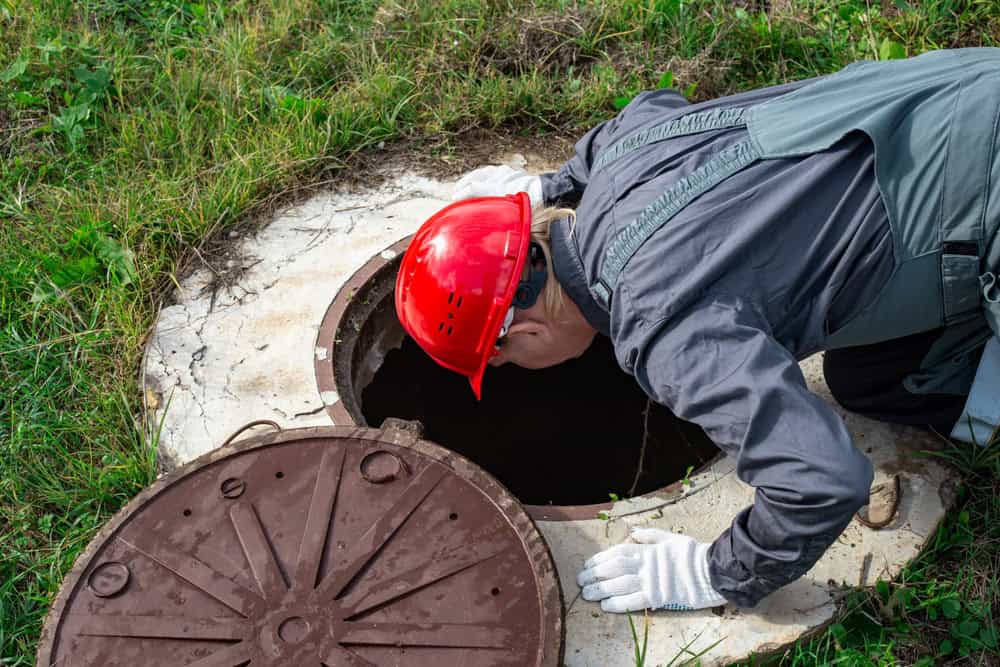 A person wearing a red hard hat and gray work clothes, representing a local septic tank service, peers into an open manhole in a grassy area, with the circular cover resting nearby.