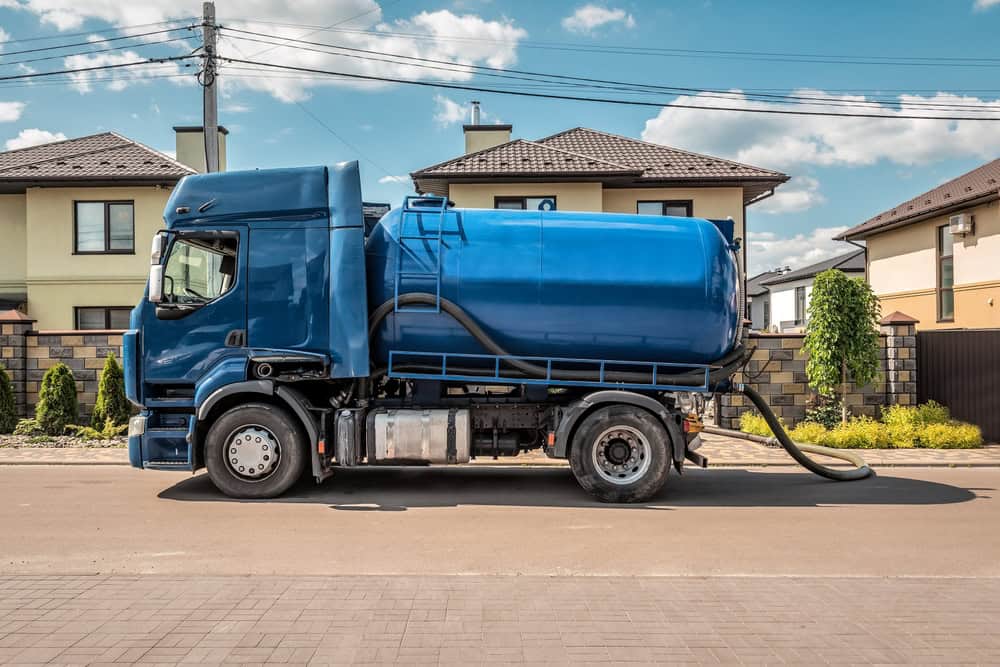 A blue septic truck, representing top-notch Septic Tank Services, is parked on a residential street in front of suburban houses with neatly maintained gardens. The sky is clear and sunny, highlighting the pristine neighborhood setting.