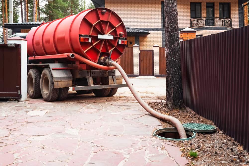 A red septic tank truck, marked for Septic Tank Services, is parked on a paved area beside a house. A hose extends into an open manhole. In the background, a wooden fence and trees provide a peaceful setting.