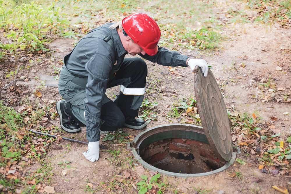 A worker in a red helmet and gray overalls is kneeling, carefully lifting the lid of a manhole outdoors among grass and fallen leaves. As part of septic tank services, they're wearing gloves while inspecting inside the open manhole.
