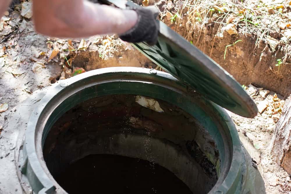 A person wearing a black glove lifts the green circular lid of an outdoor septic tank, revealing the dark interior. Dry leaves and soil surround the opening—a typical scene for septic tank services Long Island professionals in NY.