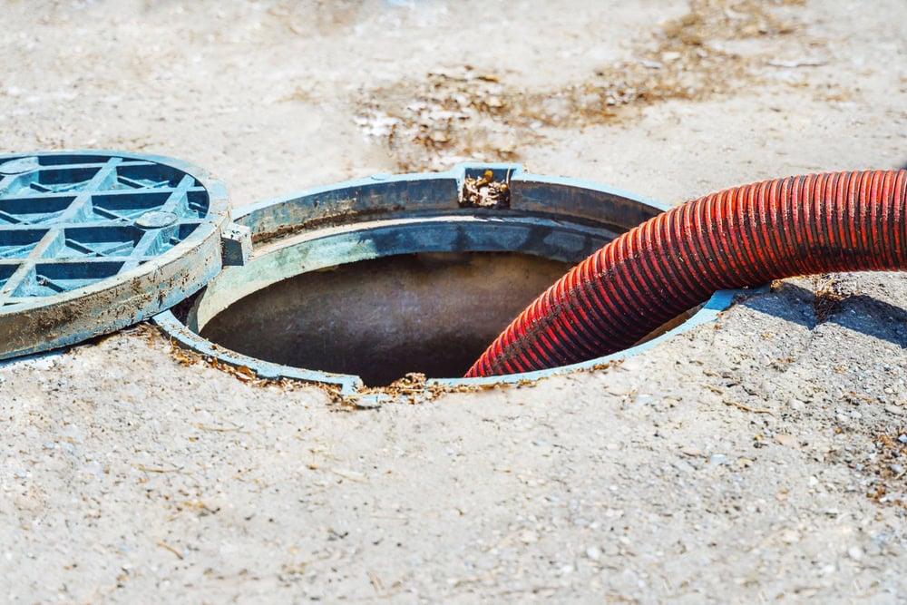 A red hose is inserted into an open manhole on a concrete surface, likely being used for cesspool services Long Island. The circular metal cover is partially removed and lies next to the opening.