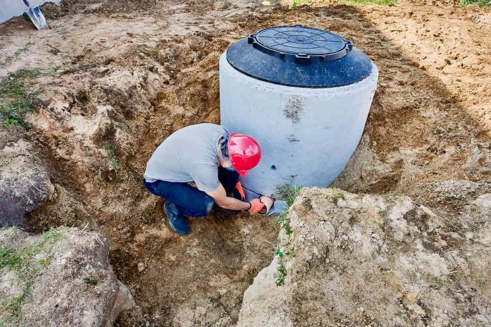 A worker wearing a red hard hat kneels beside a large, partially buried concrete septic tank, performing maintenance or installation work in a dirt excavation site—exemplifying expert septic tank services Long Island, NY.