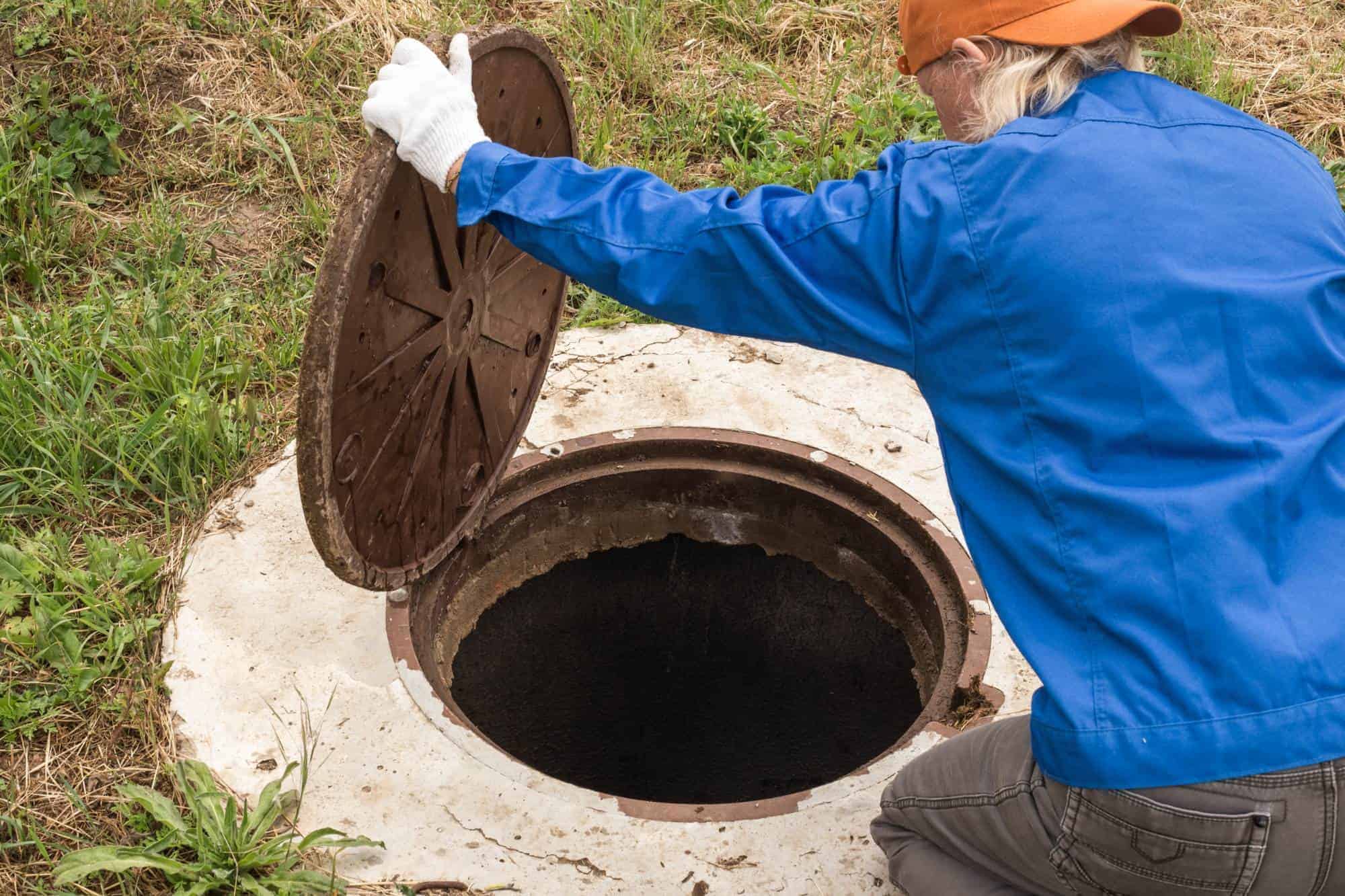 A person in a blue shirt, orange cap, and white gloves lifts the metal lid of an outdoor septic tank or manhole, revealing a dark, circular opening surrounded by grass.
