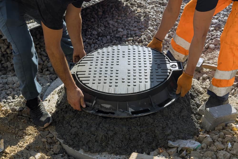 Two workers lower a round manhole cover onto a concrete base surrounded by gravel. One worker wears orange pants and gloves; the other wears dark pants.