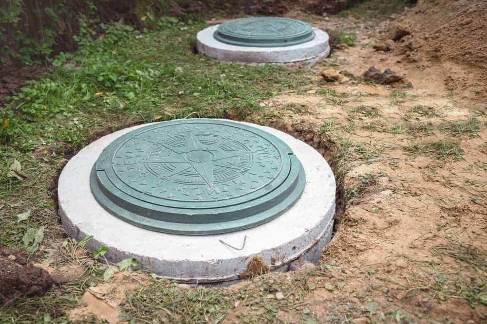 Two green septic tank covers set in concrete are embedded in the ground, surrounded by grass, dirt, and a bit of vegetation. The covers have a decorative compass design on top.