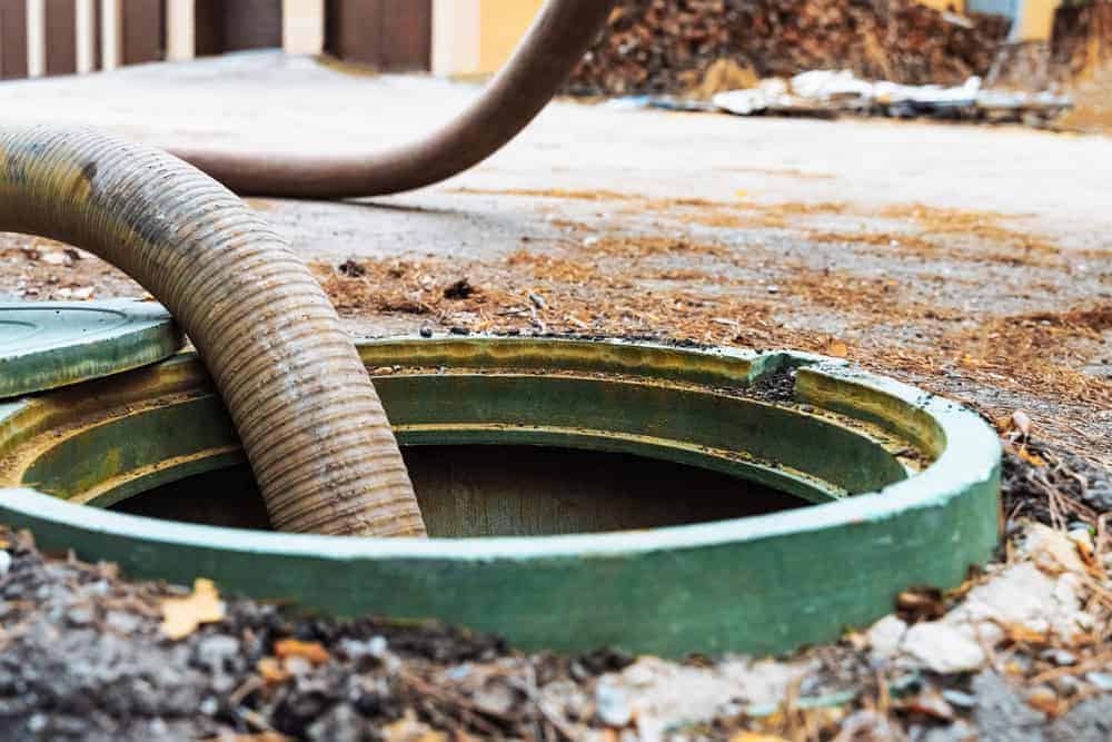 A large hose inserted into an open septic tank outdoors, with dirt and leaves scattered around the green tank cover. The background is blurred, showing piles of debris and building structures.