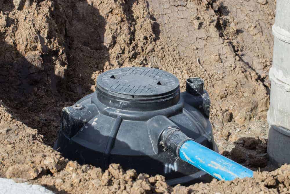 A black plastic septic tank with a round lid is partially buried in soil, with a blue pipe connected to its side and surrounding dirt visible.
